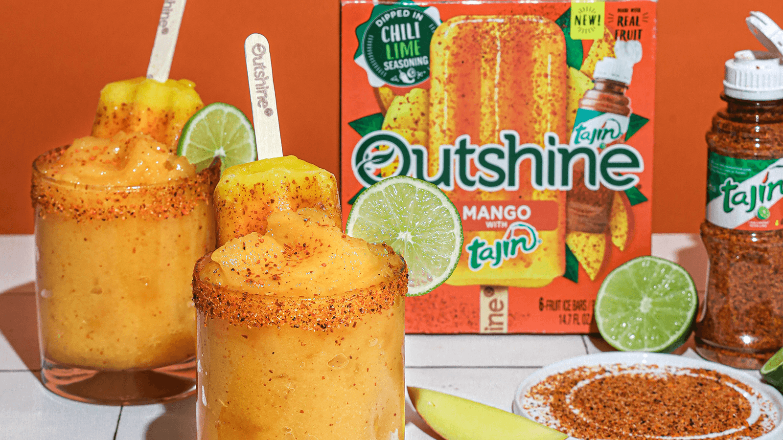 Table with Outshine Mango Tajin Frozen Fruit Bars, glasses filled with Mango Tajin Frozen Mocktails, and a plate with tajin on it.